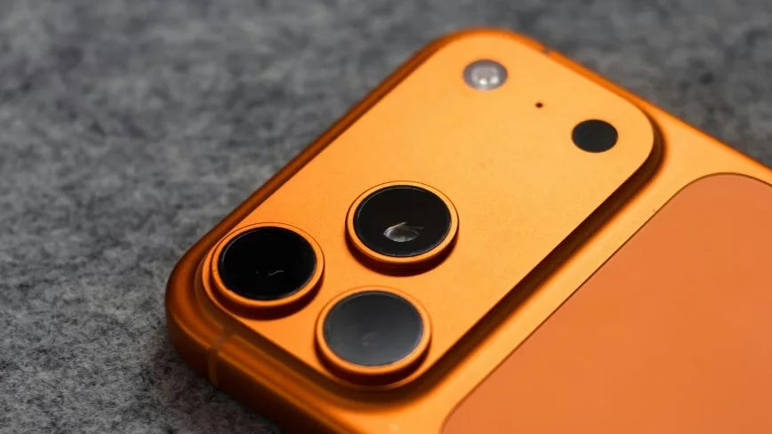 Крупний план верхнього кута помаранчевого смартфона з трьома камерами на сірій текстурованій поверхні.