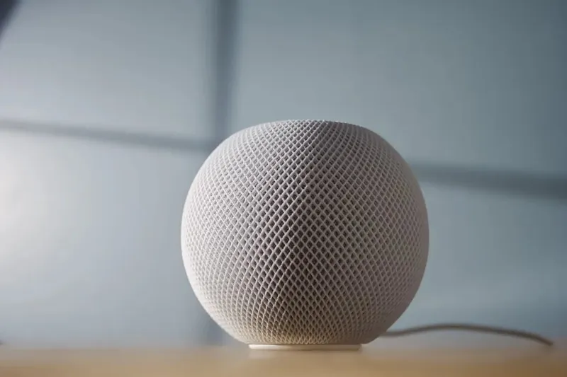 Сферичний динамік Apple HomePod білого кольору з характерною сіткою розташований на дерев'яній поверхні перед вікном.