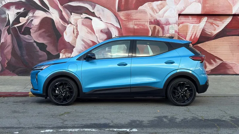 Блакитний автомобіль Chevrolet Bolt EV у боковому профілі на тлі цегляної стіни з художнім зображенням великих рожевих квітів.