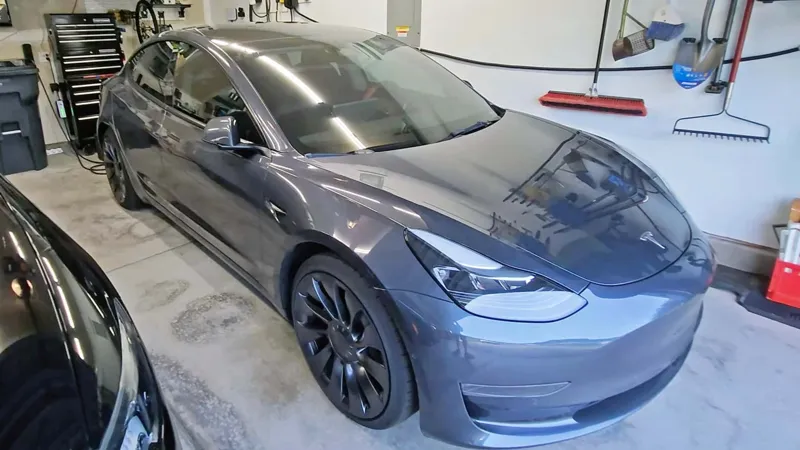 Темно-сіра Tesla Model 3 стоїть у світлому автосервісі поруч із стелажем для інструментів та побутовими речами.