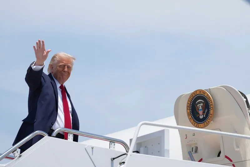 Дональд Трамп махає рукою з палуби корабля, на якому розміщено герб президента Сполучених Штатів Америки.