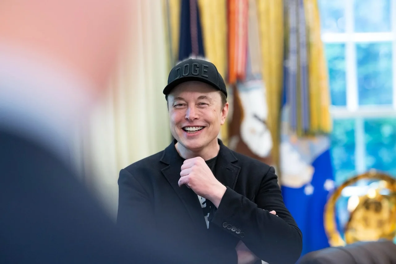 Elon Musk speaking at an official venue with corporate logos in the background.