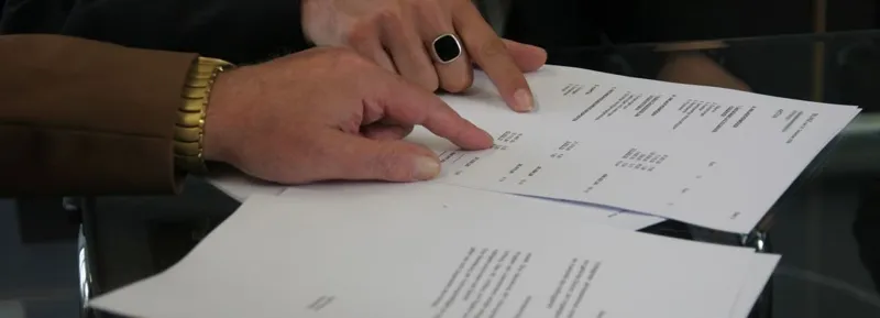 Бізнесменські руки в ділових манжетах переглядають та підписують фінансові документи на глянцевому скляному столі.