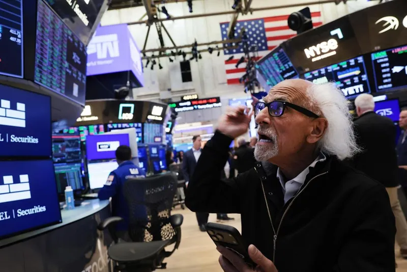 Бізнесмен із білим волоссям стоїть на торговій площі NYSE, уважно дивлячись у свій смартфон серед великих фінансових графіків та екранів.