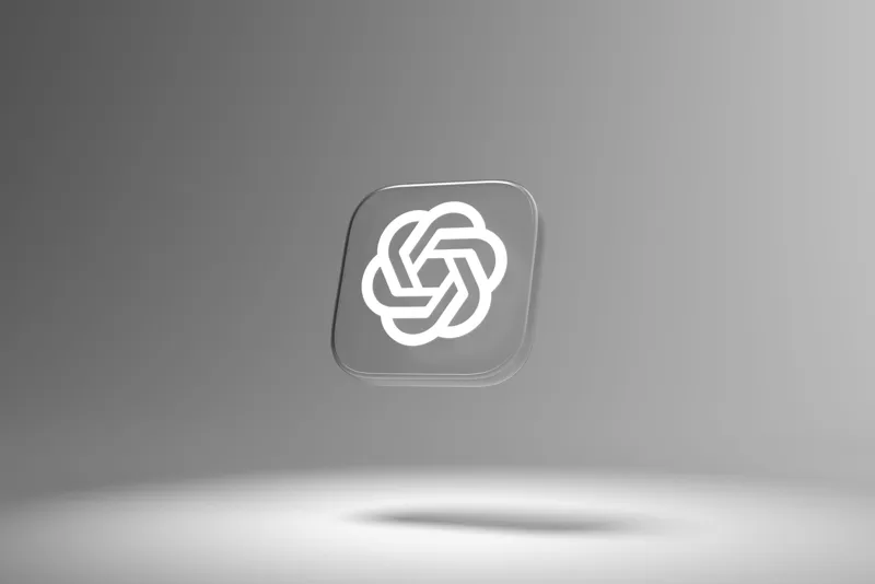 Скляна іконка з білим геометричним логотипом OpenAI, що висить у центрі перед однотонним сірим фоном із м'яким світлом знизу.