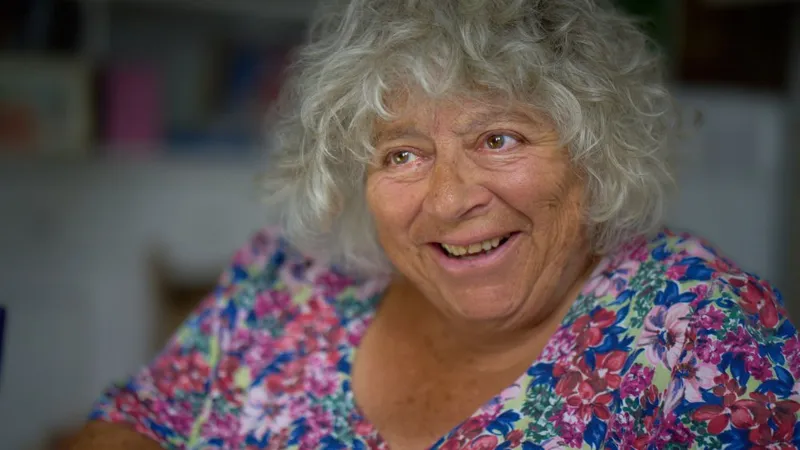 Miriam Margolyes з густим сивим волоссям у квітчастій блузці усміхається, дивлячись вбік під час церемонії.