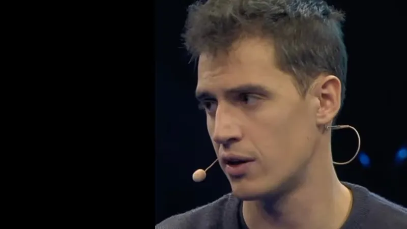 Чоловік із коротким волоссям у сірому светрі говорить у мікрофон під час виступу на сцені перед темним фоном.