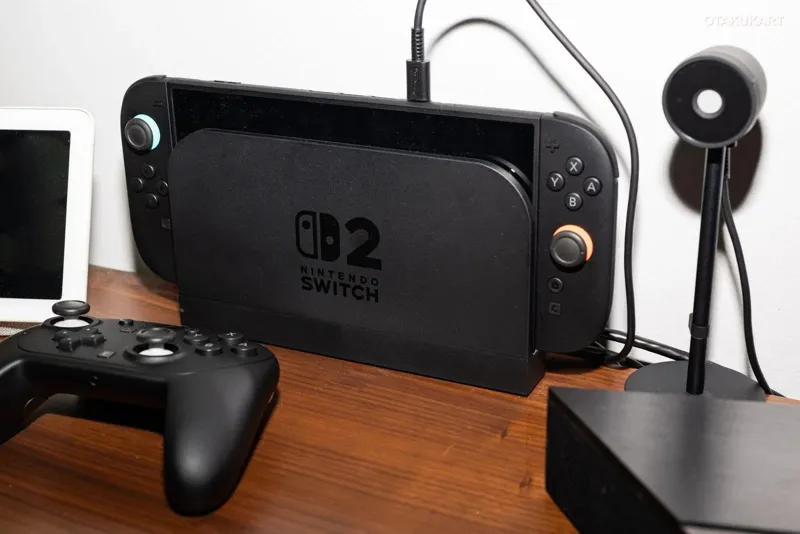 Чорна ігрова консоль Nintendo Switch з написом Switch 2 стоїть на дерев’яному столі поруч із геймпадом DualShock та мікрофоном.