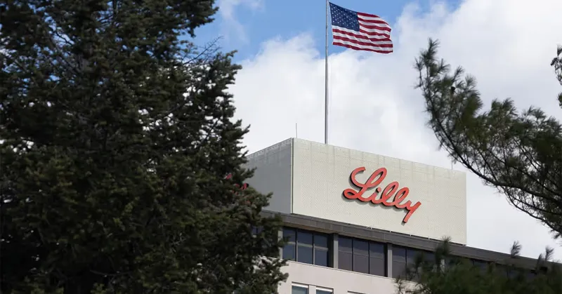 Корпоративна будівля компанії Eli Lilly з великою назвою на фасаді та американським прапором, що майорить над нею під чистим небом.