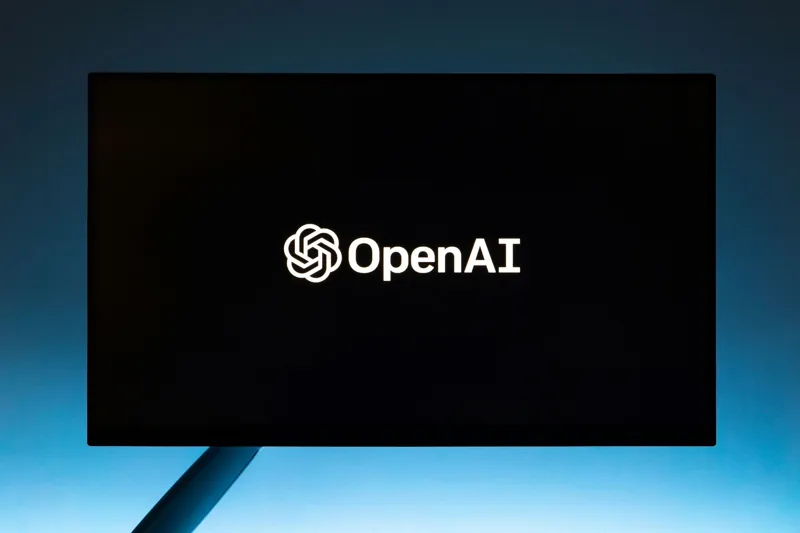 Великий чорний екран демонструє білий логотип та назву компанії OpenAI на тлі яскравого блакитного неба.