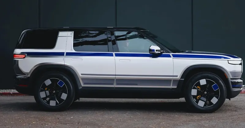 Білий електромобіль Rivian із синьо-сірими смугами на кузові та великими чорними дисками стоїть перед темною панельною стіною.