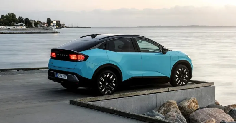 Блакитний електромобіль Nissan Ariya стоїть на бетонному пірсі біля морського берега під хмарним небом.