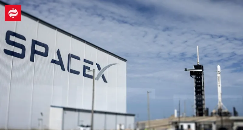 Велика біла будівля із логотипом SpaceX поруч із ракетною установкою під блакитним небом з легкими хмарами.