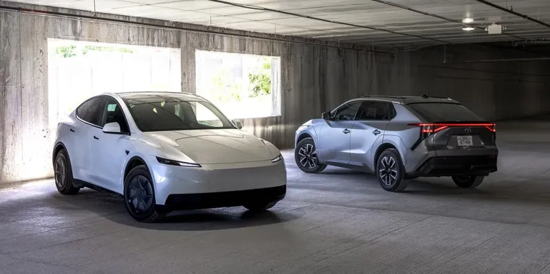 Білий кросовер Tesla Model Y та сріблястий Toyota bZ4X стоять у бетонному гаражі біля великих вікон, що пропускають світло.