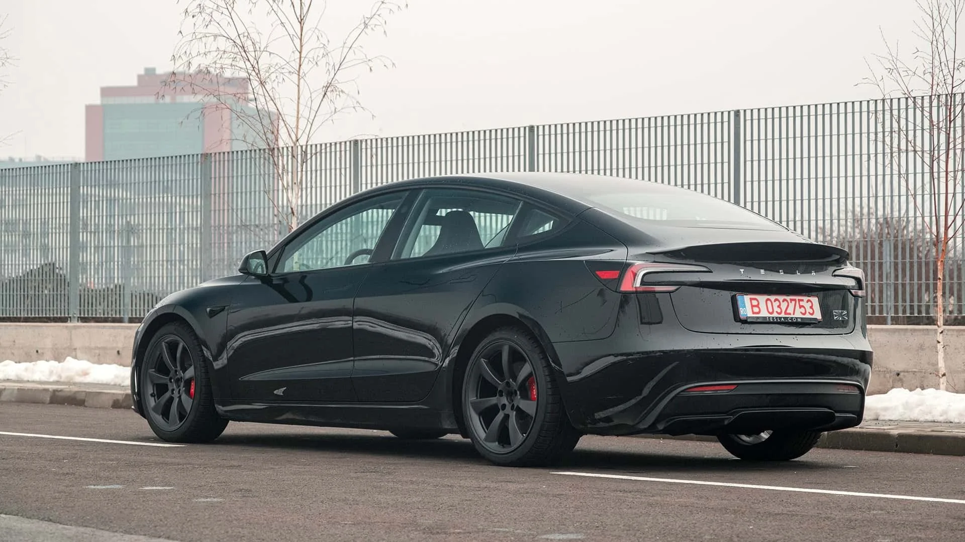 Чорний електромобіль Tesla стоїть на асфальті біля огорожі з промисловими будівлями у міському пейзажі.
