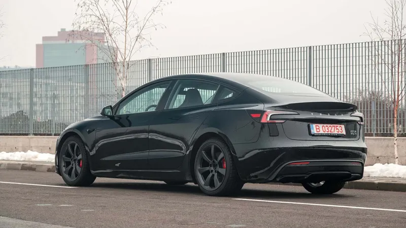 Чорний електромобіль Tesla стоїть на асфальті біля огорожі з промисловими будівлями у міському пейзажі.