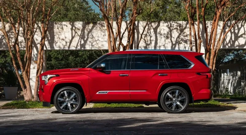 Червоний позашляховик Toyota Sequoia у профільовому виді на фоні тонких дерев і світлої стіни під відкритим небом.