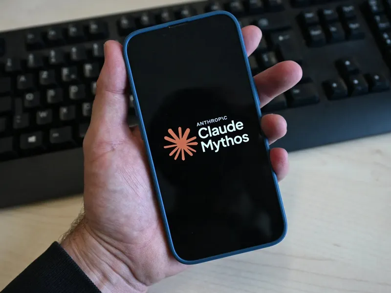 Рука тримає смартфон із логотипом Anthropic Claude Mythos на екрані перед чорною клавіатурою та дерев’яним столом.