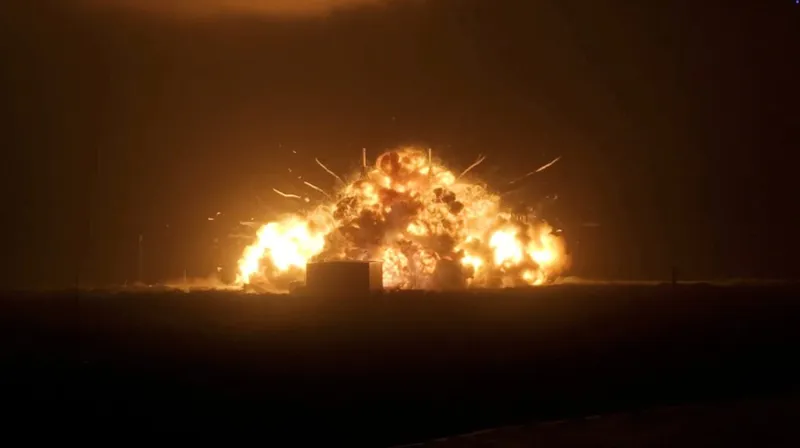 Масштабний вогонь і вибух освітлюють темне поле, де руйнується велика конструкція на стартовому майданчику.