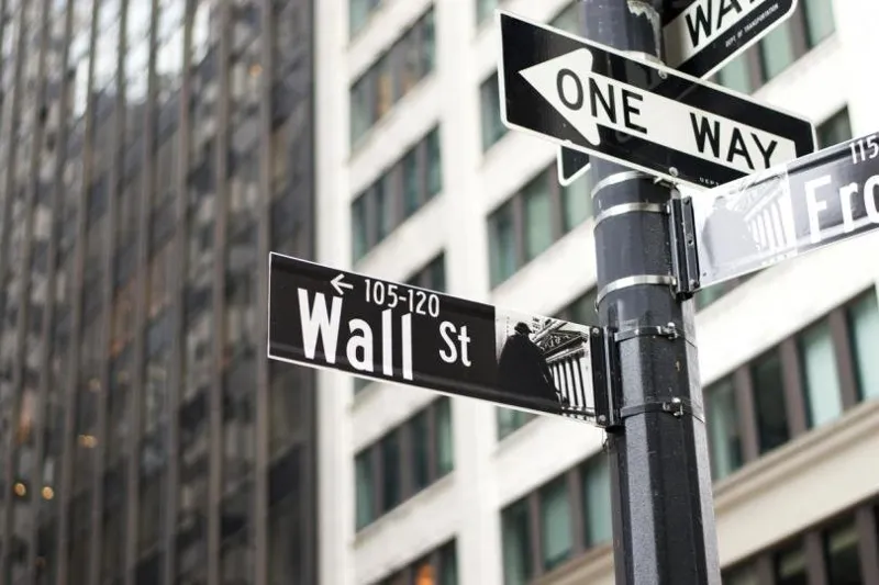 Вуличний покажчик з написом Wall St та табличка One Way на фоні багатоповерхових скляних будівель у Нью-Йорку.